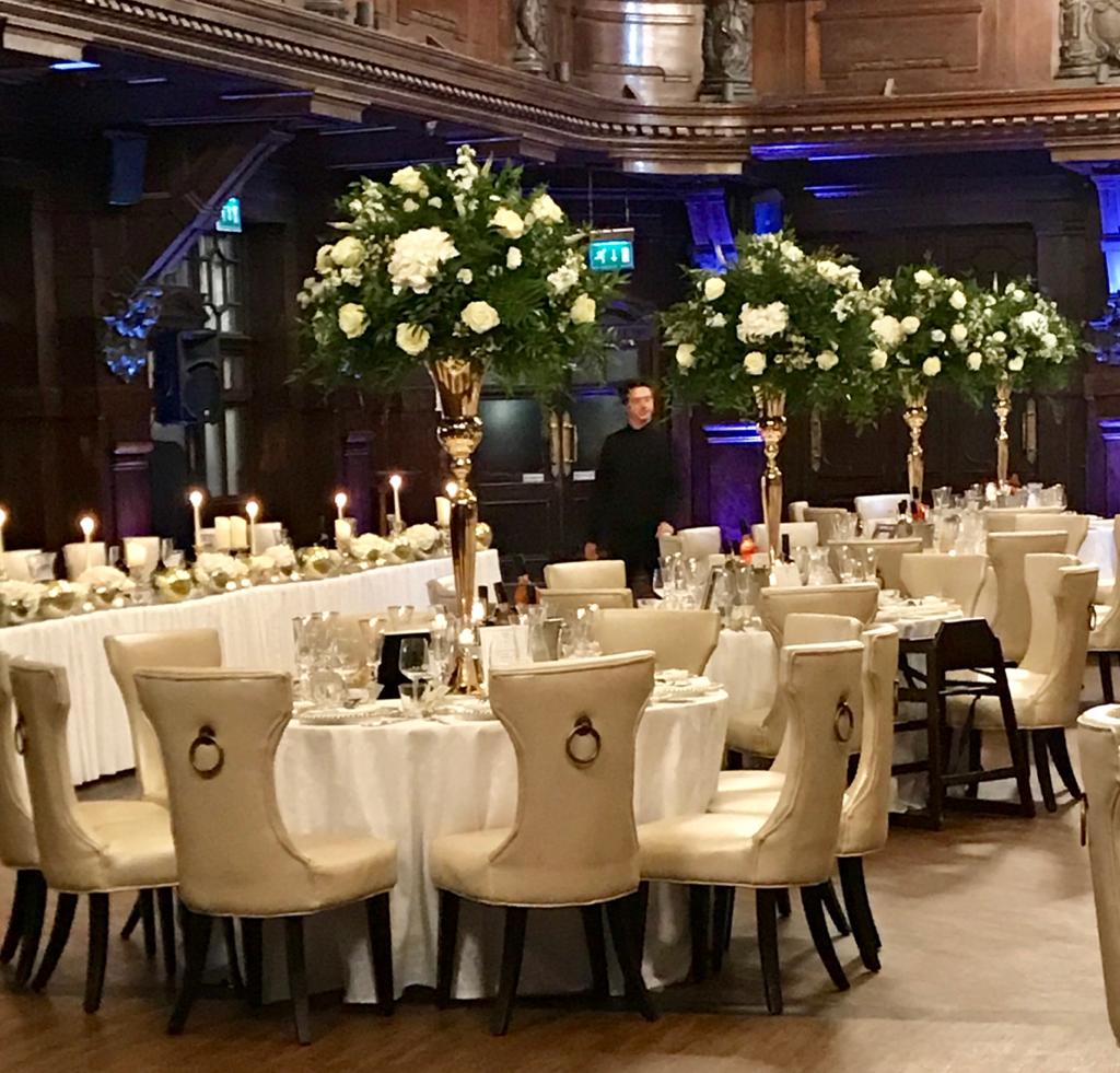 A man in a black suit stands in a room with tables and chairs set up for a wedding reception