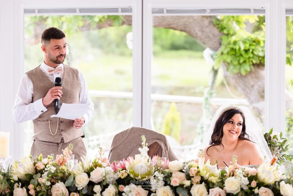 A bride and groom sitting at a table with flowers and a man giving a speech